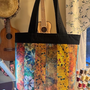 Puede incluir: Un bolso tote colorido con un estampado floral y de hojas en tonos azules, amarillos y rojos. El bolso tiene asas negras y una base negra. El fondo incluye instrumentos musicales y suministros de costura.