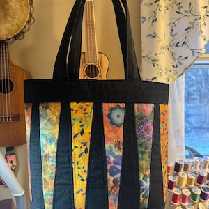 Puede incluir: Bolso tote negro con paneles verticales coloridos. Los paneles presentan diseños florales y abstractos en tonos amarillos, naranjas y morados. El bolso tiene asas negras y una base negra, ideal para llevar objetos.