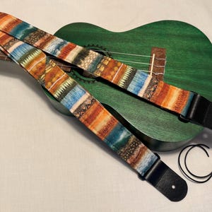 Może przedstawiać: Kolorowy pasek do ukulele z wzorem w odcieniach pomarańczu, błękitu i brązu. Pasek ma czarne skórzane końcówki i jest ułożony na zielonym ukulele. Pasek ma szerokość około 5 cm.