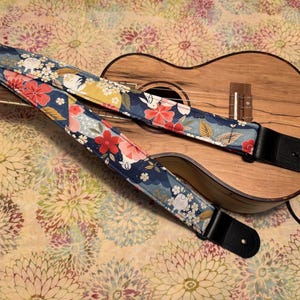 Może przedstawiać: Ukulele z paskiem w kwiatowy wzór. Pasek jest granatowy z czerwonymi, białymi i żółtymi kwiatami. Ukulele jest wykonane z drewna i ma naturalne wykończenie. Pasek jest wykonany z tkaniny i skóry.