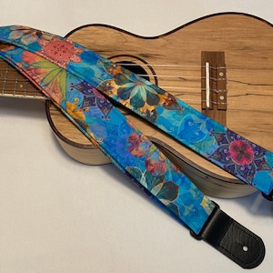 Puede incluir: Una correa de ukelele azul con un patrón floral. La correa tiene una hebilla ajustable negra y un extremo de cuero negro.
