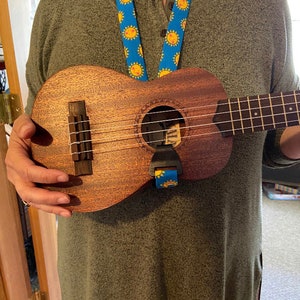 May include: A brown ukulele with a blue and yellow strap. The ukulele has a sound hole with a white ring around it. The strap has a sun design.