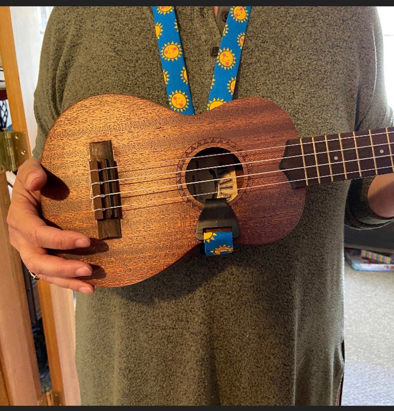 May include: A brown ukulele with a blue and yellow strap. The ukulele has a sound hole with a white ring around it. The strap has a sun design.