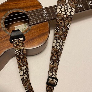 May include: A brown ukulele strap with a white floral pattern. The strap has a black buckle and is attached to a ukulele.