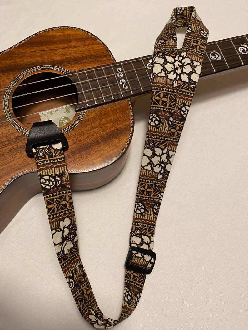May include: A brown ukulele strap with a white floral pattern. The strap has a black buckle and is attached to a ukulele.