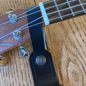May include: A black ukulele strap with a floral pattern and a silver button. The strap is attached to a ukulele with white strings.