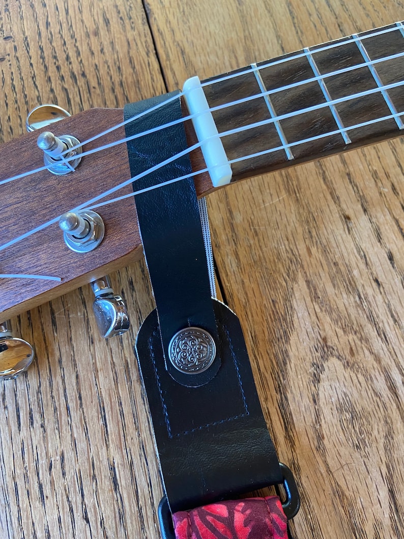 May include: A black ukulele strap with a floral pattern and a silver button. The strap is attached to a ukulele with white strings.