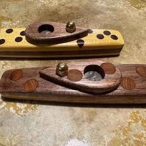 Pode incluir: Dois descansos de charuto de madeira. Um é de cor clara com pontos marrons escuros e um detalhe marrom escuro. O outro é marrom escuro. Ambos têm um descanso em forma de gota com uma porca de cor latão e um orifício circular.