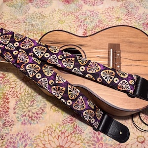 Puede incluir: Un ukelele descansa debajo de una correa de guitarra estampada. La correa presenta un fondo morado oscuro con un diseño repetido de formas amarillas y blancas. Los extremos de cuero negro completan la correa.