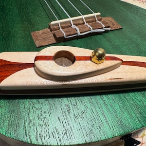 Pode incluir: Close-up de um ukulele de madeira com acabamento verde. A ponte e a pestana são visíveis, juntamente com um acessório de madeira feito sob medida com um botão de latão. O acessório tem uma base de madeira clara com detalhes em madeira escura e vermelha.