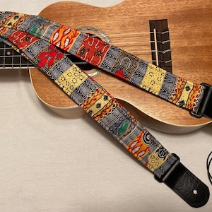 May include: A colourful patterned guitar strap with black leather ends. The strap is lying on top of a ukulele.