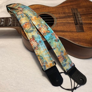 May include: A ukulele strap featuring a sea turtle design in blue, orange, and green. The strap has black leather ends and is displayed on a wooden ukulele.