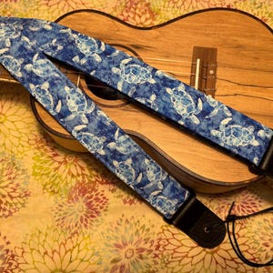 Może przedstawiać: Niebieski pasek do ukulele z wzorem białych żółwi morskich. Pasek jest wykonany z tkaniny i ma czarne skórzane końce. Ukulele jest brązowe, a tło ma kwiatowy wzór.