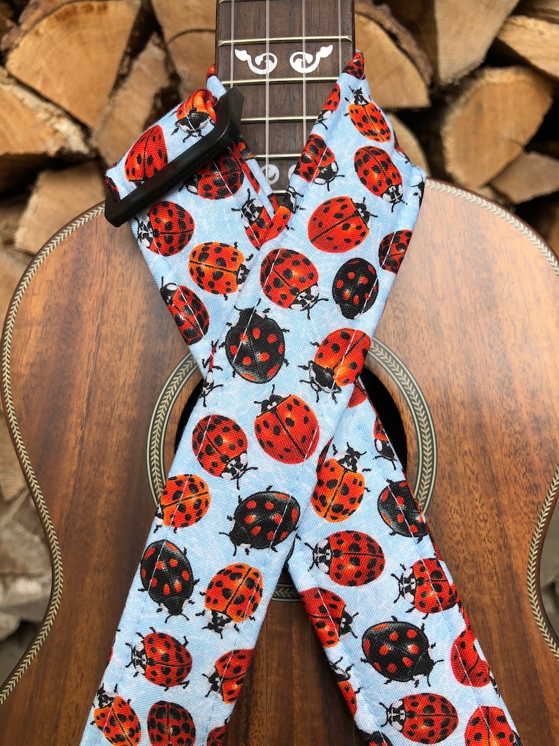 May include: A ukulele strap with a light blue background and a repeating pattern of red and black ladybirds.