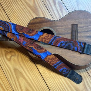 Może przedstawiać: Wzorzysty pasek do ukulele z niebieskim, brązowym i pomarańczowym wzorem, spoczywający na drewnianym ukulele. Pasek ma czarne skórzane końce i różany detal. Ukulele jest brązowe i ma otwór rezonansowy.