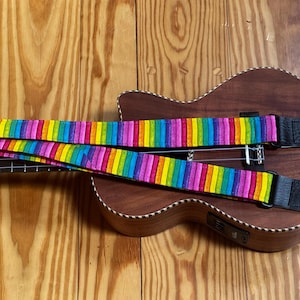 May include: A vibrant rainbow-striped ukulele strap featuring vertical bands of red, orange, yellow, green, blue, and purple. The strap includes black leather ends with pre-cut holes for attachment, resting across the body of a wooden ukulele.