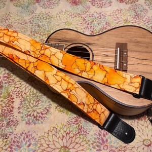 May include: A ukulele with a patterned strap. The strap features an abstract design in shades of orange and yellow, with black leather ends. The ukulele is made of wood and rests on a floral patterned surface.