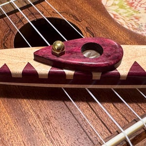 Pode incluir: Close-up de uma ponte de ukulele com um design de madeira decorativo. A ponte apresenta uma base de madeira clara com detalhes roxos e uma peça superior roxa escura. Um parafuso de metal dourado é visível. As cordas do ukulele são brancas.
