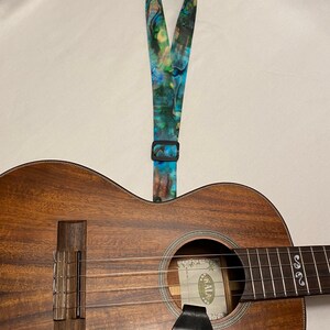 May include: A brown ukulele with a blue and green patterned strap. The ukulele has a sound hole with a label that says "Kala".