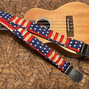 May include: A ukulele with a red, white, and blue American flag patterned guitar strap. The strap is made of fabric and has a leather end. The ukulele is on a background of music sheets.