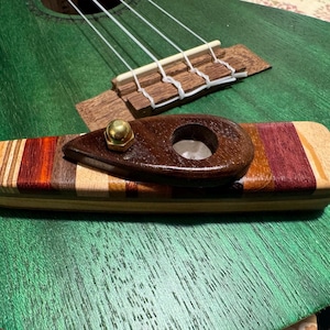Pode incluir: Um close-up de um ukulele verde com uma ponte e cordas de madeira. Um capo de madeira com uma porca de latão está no braço. O capo é feito de várias tonalidades de madeira, incluindo vermelho, castanho e bege.