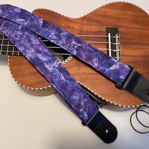May include: Purple and white patterned guitar strap with black leather ends. The strap is on top of a brown ukulele.