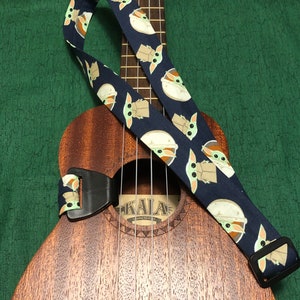 May include: A dark blue ukulele strap with a repeating pattern of a green alien character. The strap is attached to a brown ukulele with the word "KALA" on the sound hole.