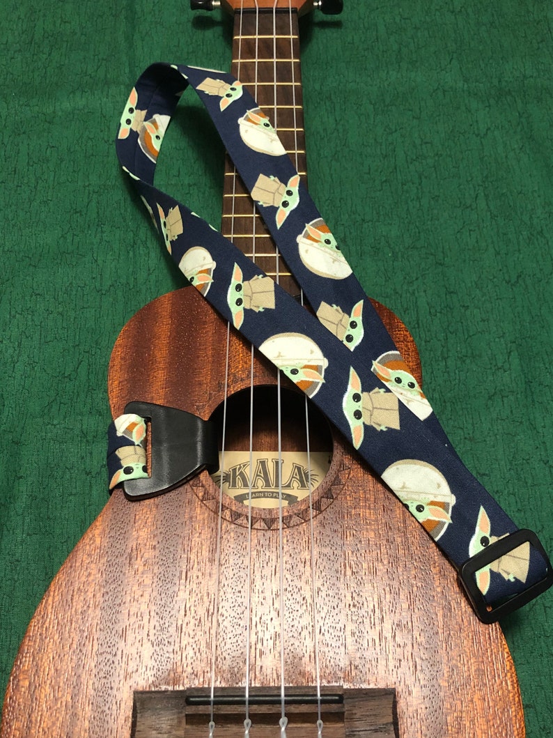 May include: A dark blue ukulele strap with a repeating pattern of a green alien character. The strap is attached to a brown ukulele with the word "KALA" on the sound hole.
