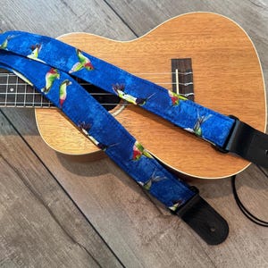 May include: A blue ukulele strap with a hummingbird pattern. The strap is made of fabric and has black leather ends. The ukulele is brown and has a wooden body. The strap is lying across the ukulele.
