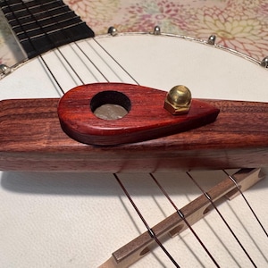 Pode incluir: Um close-up da ponte e do esticador de um banjo. A ponte é feita de madeira clara, e o esticador é de madeira castanho-avermelhada escura com uma porca de latão. As cordas do banjo são visíveis, juntamente com a pele branca.