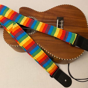 Może przedstawiać: Pasek do gitary w paski tęczowe z czarnymi skórzanymi końcówkami i czarnym regulowanym paskiem. Pasek leży na drewnianym ukulele.