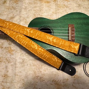 May include: A green ukulele with a mustard yellow strap featuring a white pattern. The strap has black leather ends and is draped across the ukulele's fretboard and body. The ukulele is resting on a patterned surface.