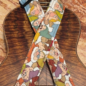 May include: A white guitar strap with a repeating pattern of the seven dwarfs from the Disney movie Snow White and the Seven Dwarfs. The strap is shown on a wooden guitar.