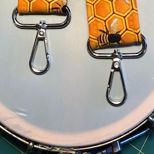 May include: Two silver swivel clasps with a honeycomb and bee pattern fabric strap. The strap is yellow and black with a repeating pattern of honeycombs and bees.