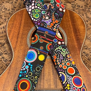 Może przedstawiać: Kolorowy pasek do gitary z czarnym tłem i wzorem kół w różnych rozmiarach i kolorach. Pasek jest skrzyżowany na drewnianym ukulele.