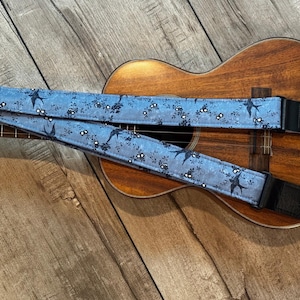 May include: A light blue guitar strap featuring a dark blue pattern of small flowers and flying swallows. The strap has black leather ends with adjustment buckles and is positioned across the body of a brown wooden acoustic ukulele.