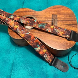 Może przedstawiać: Brązowy pasek do ukulele z kolorowym wzorem piór. Pasek jest przymocowany do ukulele z drewnianym korpusem.