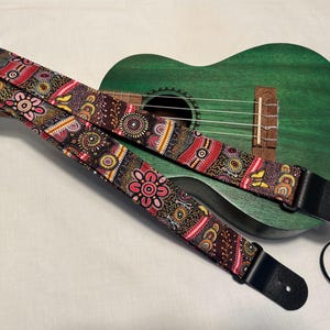 Może przedstawiać: Pasek do gitary z kolorowym wzorem i czarnymi skórzanymi końcówkami. Pasek jest przymocowany do zielonego ukulele.