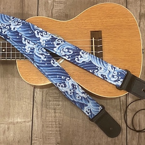 Puede incluir: Una correa de ukelele azul y blanca con un patrón de olas. La correa está hecha de tela y tiene un extremo de cuero negro.