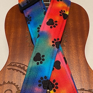 Może przedstawiać: Kolorowy pasek do ukulele z wzorem tie-dye i czarnymi odciskami łap. Pasek jest wykonany z tkaniny i ma czarną regulowaną klamrę.