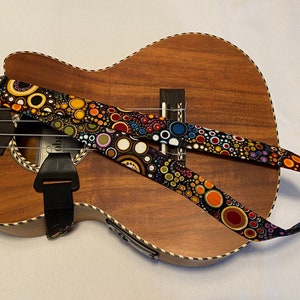Może przedstawiać: Brązowy ukulele z czarno-białym paskiem w kolorowe koła. Pasek jest przymocowany do ukulele czarnym paskiem skórzanym.