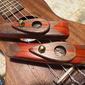 Pode incluir: Duas pontes de madeira para ukulele com uma combinação de cores marrom escuro e vermelho. Cada ponte tem um orifício circular e uma porca de cor latão. As pontes estão no braço de um ukulele.
