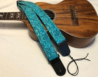 May include: A teal blue ukulele strap with a swirling wave pattern. The strap has black leather ends and a black adjustable strap.