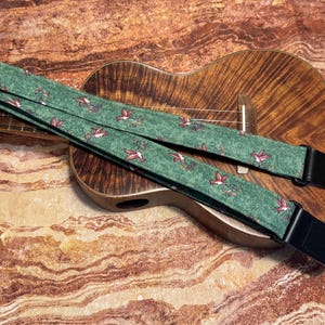 Może przedstawiać: Zielony pasek do gitary z wzorem kolibra i czarnymi skórzanymi końcówkami. Pasek leży na drewnianej gitarze.