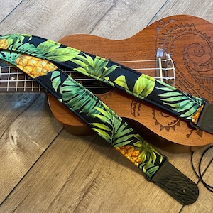 May include: A black ukulele strap with a green and yellow pineapple and leaf pattern. The strap is on a ukulele with a carved design.
