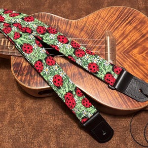 May include: A black guitar strap with a red and black ladybug pattern on a green background. The strap is attached to a ukulele with a brown wooden body.