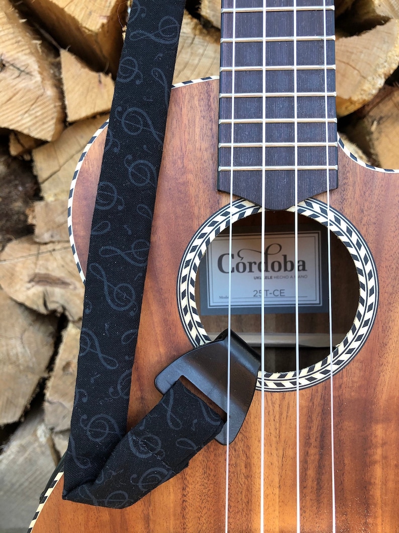 May include: A brown wooden ukulele with a black and white patterned soundhole. The ukulele has a black strap with a musical note pattern. The ukulele is labeled "Cordoba" and "25T-CE".