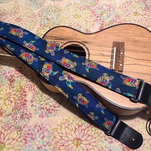 May include: A blue ukulele strap featuring a pattern of colourful turtles, lying on a wooden ukulele. The strap has black leather ends and is decorated with vibrant, multi-coloured turtles. The ukulele has a natural wood finish.