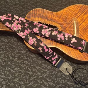 May include: A black ukulele strap with a pink floral pattern. The strap is attached to a ukulele with a brown wooden body.