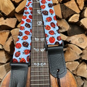 May include: A ukulele with a black leather strap and a blue and white ladybug patterned strap. The ukulele has a wooden body and silver-coloured tuning pegs. The ukulele is labelled "KALA" on the body.